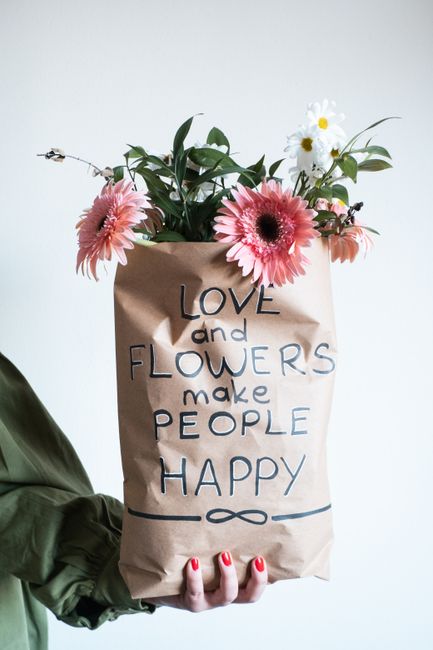 A person holding a brown paper gift bag filled with an arrangement of fresh flowers, including pink gerbera daisies, white daisies with yellow centers, and green foliage. The bag features handwritten 