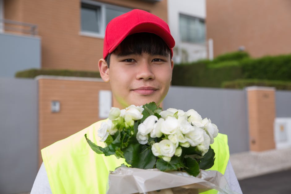 A young male delivery courier wearing a bright yellow vest and a red cap is holding a bouquet of white roses and green foliage wrapped in white paper. The flowers appear fresh with slightly open petal