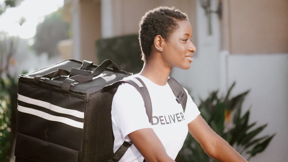 A female flower delivery courier with short, curly hair and a focused expression standing outdoors near a residential area. She is wearing a white T-shirt with the word 'DELIVERY' partially visible an