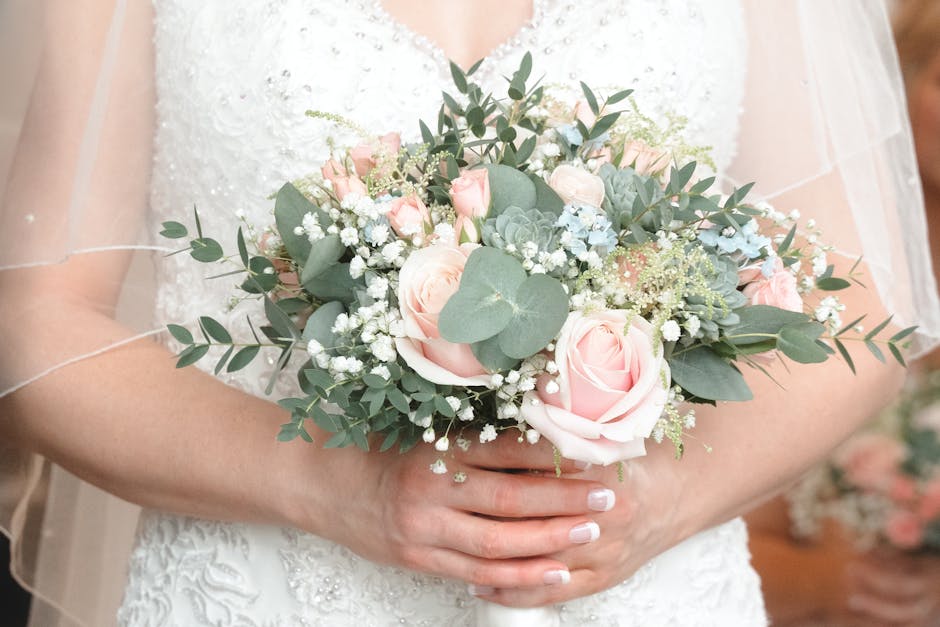 A bride holds a bouquet arrangement consisting of soft pink roses, white baby's breath, and eucalyptus leaves with rounded, silvery-green foliage. The flowers are fresh, with delicate petals and sturd