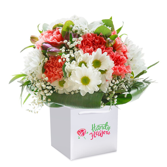 A generously rounded floral bouquet, expertly arranged and displayed upright in a pristine, square white gift bag, captures the cosmopolitan freshness of Gospel Oak in full bloom. The arrangement centres on lush, ruffled carnations in soft coral, gentle peach, and bright scarlet-red, each blossom brimming with intricate texture and vitality akin to spring's first burst along Gospel Oak's leafy paths. Between them, clusters of white daisy-like blooms with brilliant yellow-green centers add spirited contrast and lift, their crisp petals gleaming with pure optimism. Wispy branches of white baby's breath weave throughout, offering softness and volume, while enigmatic, closed buds tinged with purple and blush extend from the left, lending depth and a sense of unfolding promise. The base of the bouquet is richly ringed by lush, deep green leaves-broad, glossy, and meticulously arranged to create a graceful, natural collar reminiscent of the foliage in nearby recreational gardens. This thoughtfully constructed bouquet, shaped to form a full, welcoming dome, speaks to the careful attention of local florists familiar with Gospel Oak's diverse tastes. The white gift bag serves as both a secure and elegant frame, featuring sturdy white ribbon handles at the top-a detail designed as much for practicality on local deliveries as for refined presentation. Set against a spotless white backdrop, the florals seem to radiate with freshness, highlighting their suitability for marking life's uplifting moments, whether that's an anniversary near Oaklands Road, a birthday on Codrington Hill, or just reaching out to someone dear in Gospel Oak with a flourish of colour and care.